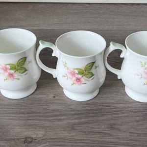 May include: Five white ceramic mugs with a floral design of pink flowers and green leaves. Each mug has a rounded base, a curved handle, and a light green accent on the handle. The mugs are arranged in a row.