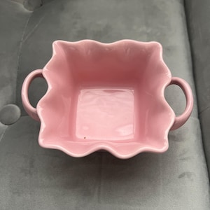 May include: A square, pink ceramic bowl with a scalloped edge and two small handles. The bowl is a soft, pastel pink colour and appears to be glazed ceramic. The bowl is on a grey surface.