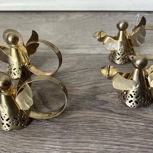 May include: Four gold-coloured angel napkin rings. Two angels are playing trumpets, and the other two are holding candle holders. The angels have wings and are attached to a circular ring. The napkin rings are made of metal.