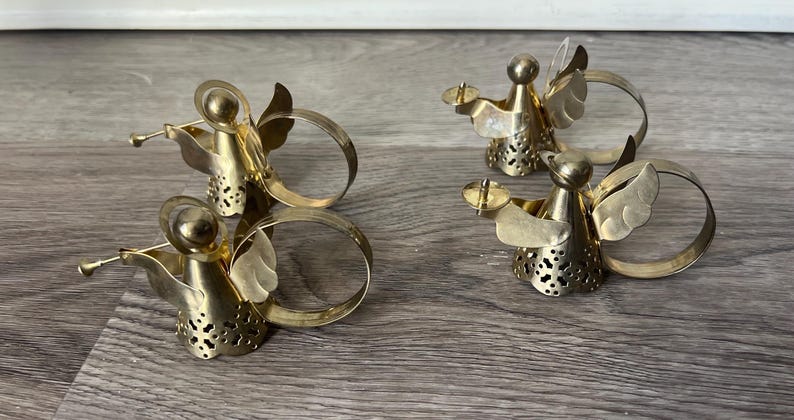 May include: Four gold-coloured angel napkin rings. Two angels are playing trumpets, and the other two are holding candle holders. The angels have wings and are attached to a circular ring. The napkin rings are made of metal.