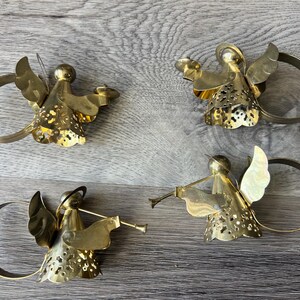 May include: Four brass angel napkin rings. Each angel has detailed wings and a halo, holding various objects, including a trumpet. The napkin rings are circular and attached to the angel's body. The angels are on a grey wooden surface.