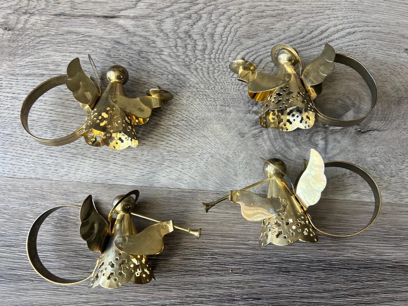 May include: Four brass angel napkin rings. Each angel has detailed wings and a halo, holding various objects, including a trumpet. The napkin rings are circular and attached to the angel's body. The angels are on a grey wooden surface.