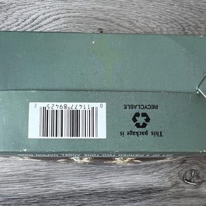 May include: A sage green box with a barcode and the text "This package is recyclable." The box is on a wooden surface. The box is for a set of pierced two-tone angel napkin rings.