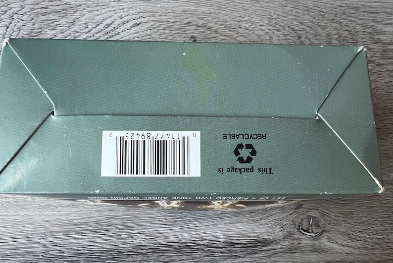 May include: A sage green box with a barcode and the text "This package is recyclable." The box is on a wooden surface. The box is for a set of pierced two-tone angel napkin rings.
