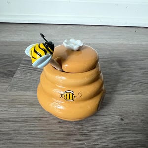 May include: A yellow ceramic honey pot shaped like a beehive with a matching lid featuring a white flower handle. A small bee is painted on the side of the pot, and a honey dipper with a bee-shaped handle rests inside the opening of the lid.