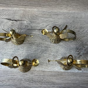 May include: Four vintage brass angel napkin rings. Each angel has wings, a trumpet, and a ring for holding a napkin. The angels are gold-coloured and rest on a wooden surface.