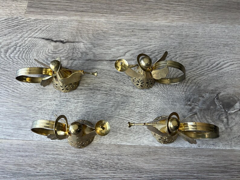 May include: Four vintage brass angel napkin rings. Each angel has wings, a trumpet, and a ring for holding a napkin. The angels are gold-coloured and rest on a wooden surface.