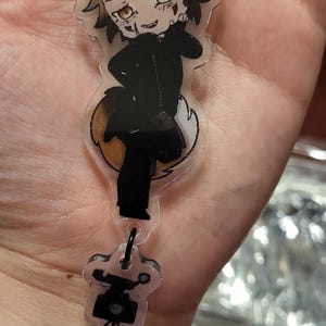 Könnte beinhalten: Ein klarer Acryl-Schlüsselanhänger mit einem cartoonartigen fuchsähnlichen Charakter mit braunen und schwarzen Details. Der Schlüsselanhänger hat einen schwarzen Verschluss und einen kleinen Anhänger am Boden. Die Figur trägt ein schwarzes Outfit.