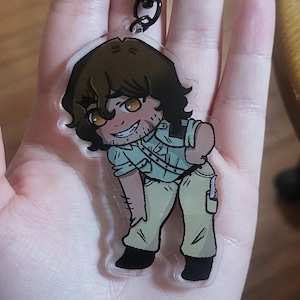 May include: A clear acrylic keychain featuring a cartoon illustration of a person with brown hair, wearing a light green shirt and tan pants. The keychain has a black heart-shaped clasp.