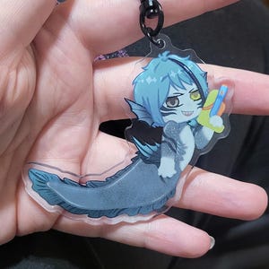 Puede incluir: Un llavero acrílico transparente con un personaje de dibujos animados con cabello azul y cola de pez. El personaje sostiene una pistola de agua colorida. El llavero tiene un cierre negro en forma de corazón.