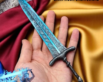 May include: A detailed miniature sword with a blue blade and ornate silver handle. The blade features intricate leaf-like patterns. The text "DARK MOON GREATSWORD" is visible in the background. The sword is held in a hand.