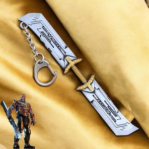 May include: A silver and gold double-edged sword keychain with a detailed design and a silver ring. The sword has a black and gold pattern on the blade.