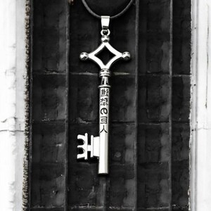 May include: Silver-colored key pendant with a diamond-shaped top and Japanese text. The key hangs from a black cord against a dark, textured background. The key's design is intricate and detailed.