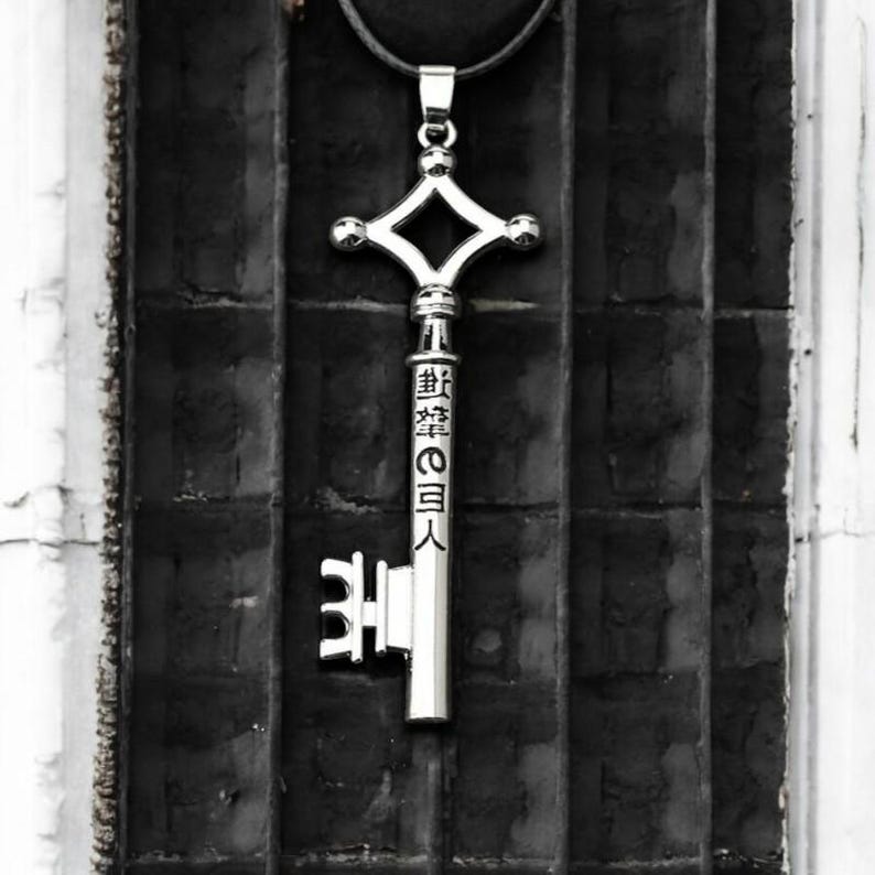 May include: Silver-colored key pendant with a diamond-shaped top and Japanese text. The key hangs from a black cord against a dark, textured background. The key's design is intricate and detailed.