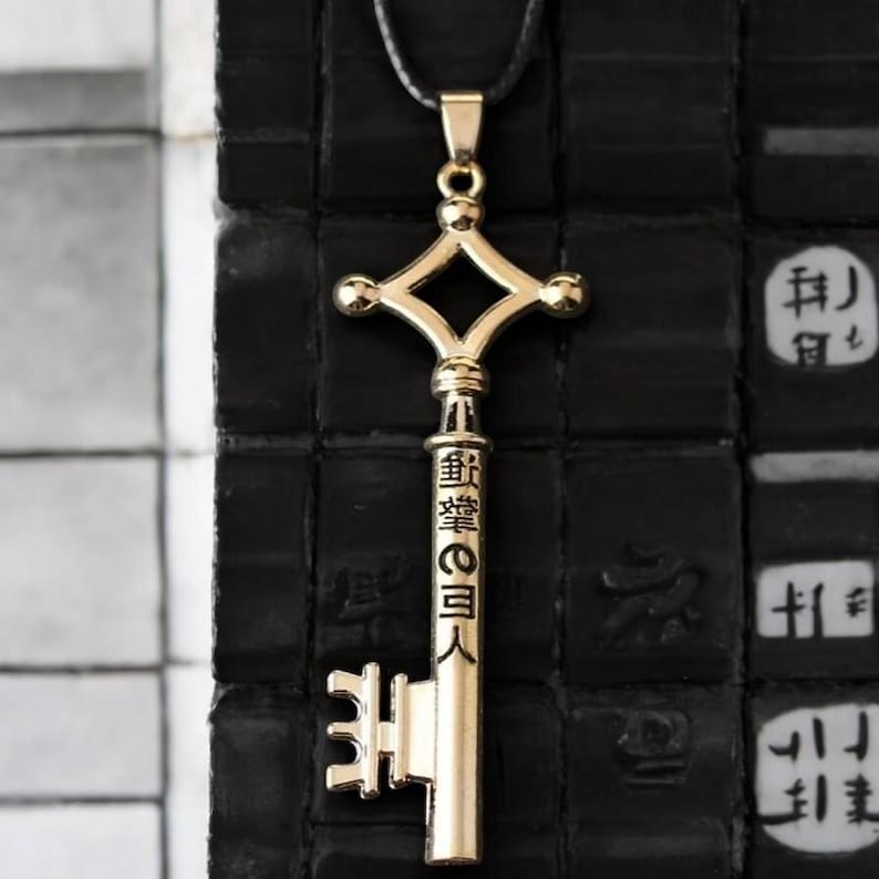 May include: A gold-colored pendant necklace featuring a key design. The key has a cross-shaped top and a detailed shaft with Japanese characters. The pendant hangs from a black cord, set against a dark background.