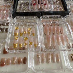May include: A collection of clear plastic containers filled with various sets of artificial nails. The nails are in different shapes and sizes, and some have gold accents or glitter.