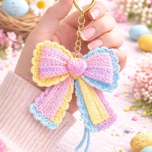 Pode incluir: Um porta-chaves de laço de crochê em tons pastel com um fecho dourado em forma de coração. O laço apresenta secções rosa, amarelas e azuis, com uma borda ondulada. Um acessório decorativo.