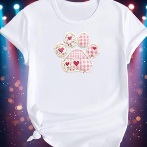 Könnte beinhalten: Weißes T-Shirt mit einem Pfotenabdruck-Design. Der Pfotenabdruck besteht aus rosa Vichy- und Blumenmustern, jedes mit einem roten Herzen. Das Design ist mit einer dekorativen Bordüre versehen.