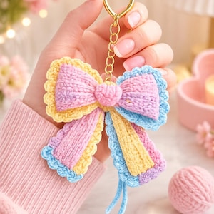 May include: A handmade crochet bow keyring featuring a pastel colour palette of soft pink, pale yellow, and light blue yarn. The bow is attached to a gold-toned metal heart-shaped ring and a short chain, with two dangling yarn tails at the bottom.