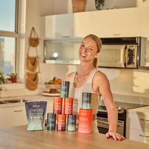 May include: A woman smiles in a kitchen, standing behind a table displaying health and wellness products. The products include cylindrical containers and a pouch, all with branding. The kitchen has white cabinets, a microwave, and a window.