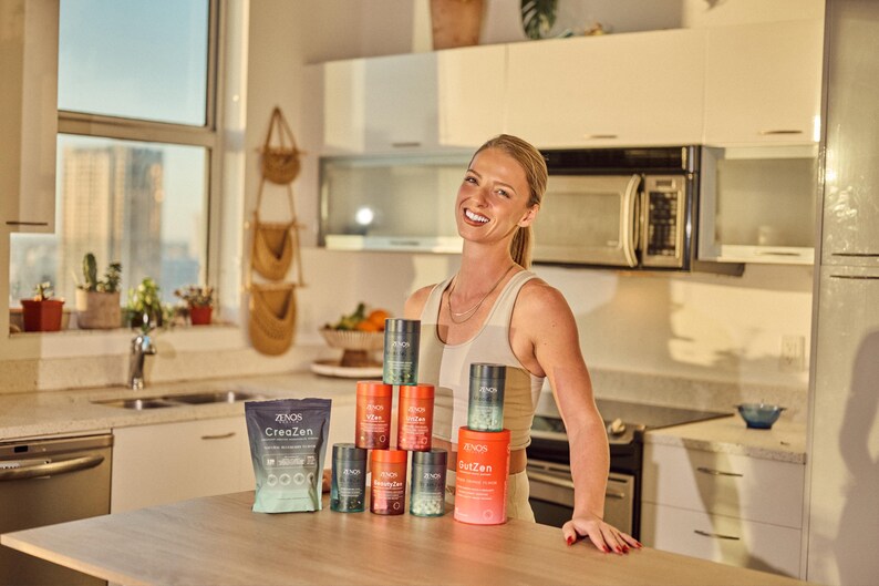 May include: A woman smiles in a kitchen, standing behind a table displaying health and wellness products. The products include cylindrical containers and a pouch, all with branding. The kitchen has white cabinets, a microwave, and a window.