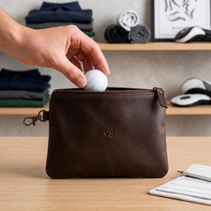 Könnte beinhalten: Eine dunkelbraune Leder-Reißverschlusstasche mit den eingeprägten Buchstaben VS auf der Vorderseite. Die rechteckige Tasche verfügt über einen seitlichen Metallclip und wird mit einem weißen Golfball mit Dellen gezeigt.