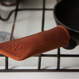May include: A brown leather pan handle cover featuring visible stitching along the edges and the embossed letters RV on the top surface. The cover is fitted over the handle of a black frying pan resting on a metal stovetop grate.