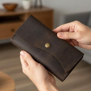 Op de afbeelding: Een donkerbruine leren portemonnee met een klepsluiting en een messing knoop. De portemonnee is rechthoekig en lijkt gemaakt van leer met een gebruikte look. De portemonnee wordt door twee handen vastgehouden.