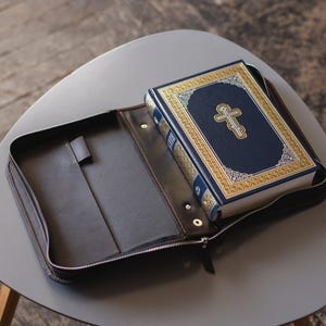 May include: A dark brown leather zipped book cover containing a blue hardback book. The book features a gold-coloured Orthodox cross on the front cover, intricate gold border detailing, and a metallic spine. The interior of the cover is dark brown.