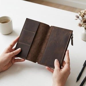 Pode incluir: Uma carteira de couro castanho escuro com orientação vertical, apresentando múltiplas ranhuras para cartões no lado esquerdo e um compartimento com fecho no lado direito, aberta para mostrar a estrutura interior e a textura do couro.