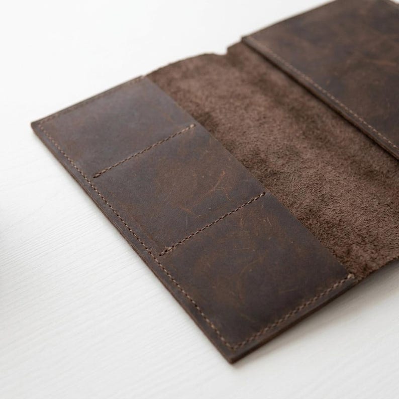 May include: A dark brown leather wallet with a matte, distressed finish. The interior features a smooth, textured surface with two distinct card slots on the left side and a larger open pocket on the right side, all held together by visible stitching.