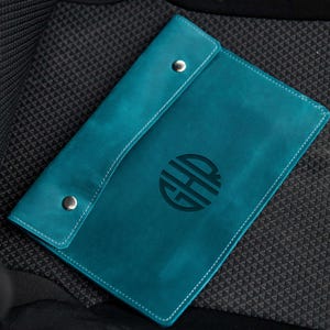 May include: Teal leather folder with a monogrammed circle in the centre. The monogram is the letters "HH" in a sans-serif font.