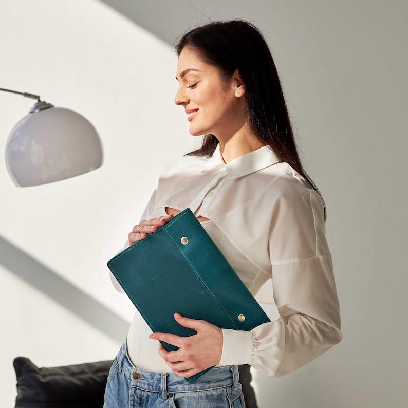 May include: A woman is holding a teal leather portfolio with silver accents. The portfolio is closed and has a smooth surface.