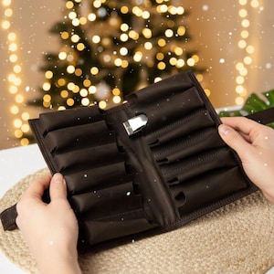 Puede incluir: Un estuche enrollable marrón oscuro, abierto, con múltiples compartimentos, sostenido abierto por manos. El estuche tiene un cierre plateado y una correa. El fondo presenta un árbol de Navidad con luces borrosas y una lluvia de puntos blancos.