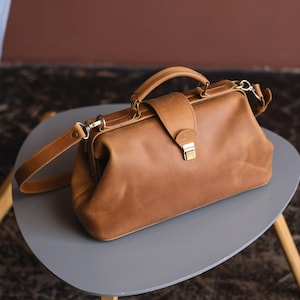 May include: A brown leather doctor's bag with a top handle and a long shoulder strap. The bag has a brass clasp and a flap over the top.