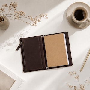 May include: A dark brown leather journal cover featuring a zippered pocket on the left side and a tan paper notebook insert on the right. The cover is open, revealing the interior structure with metal binding rings visible along the center spine.
