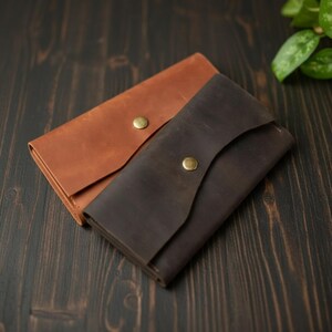 May include: Two rectangular leather wallets, one in a light tan colour and one in a dark brown colour. Each wallet features a single flap closure secured with a centred brass-coloured metal snap button. The leather has a matte, smooth finish.
