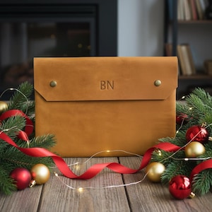Puede incluir: Funda para portátil de cuero color canela con las iniciales "BN" grabadas en la parte delantera. La funda tiene un cierre de solapa asegurado con dos remaches de latón. Decoraciones festivas con adornos rojos y dorados, cinta roja y ramas de pino.