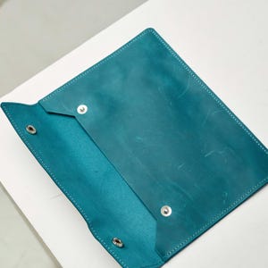 May include: A teal leather folder with a flap closure and metal poppers. The folder is open and the inside is visible.