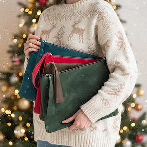 May include: A collection of leather clutch bags in various colors, including teal, red, olive green, and brown. Each bag has a zipper closure and some feature a wrist strap or tassel. The bags are held in front of a festive Christmas tree.