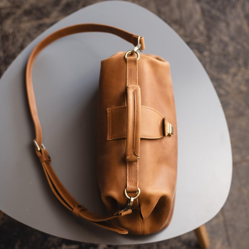May include: A brown leather duffel bag with a long strap and a top handle. The bag has a gold buckle and a gold zipper pull.