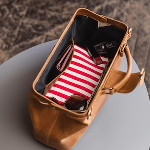 May include: A brown leather doctor bag with a red and white striped fabric lining. The bag is open and contains a wallet, sunglasses, and makeup.