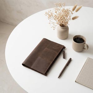 May include: A dark brown leather zippered portfolio case with a front pocket, accompanied by a matching brown pen, a beige ceramic mug filled with dark coffee, a small beige vase containing dried flowers, and a light gray notebook.