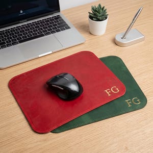 Könnte beinhalten: Ein rotes und ein dunkelgrünes rechteckiges Mauspad aus Leder, jeweils mit den goldgeprägten Initialen FG versehen. Eine schwarze Computermaus liegt auf dem roten Pad. Die Pads liegen auf einer Holzoberfläche neben einem Laptop und einer Pflanze.