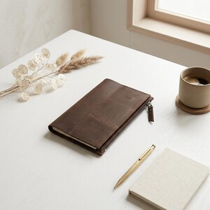 May include: A rectangular dark brown leather notebook cover featuring a side zipper closure with a matching leather pull tab. The cover has visible stitching along the edges and a smooth matte finish, designed to hold a notebook securely inside.