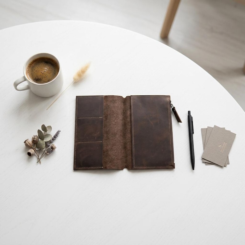 May include: A dark brown leather wallet with a zippered compartment and multiple card slots, accompanied by a black pen, a stack of grey business cards, a white mug filled with coffee, and a small sprig of dried flowers and seed pods.