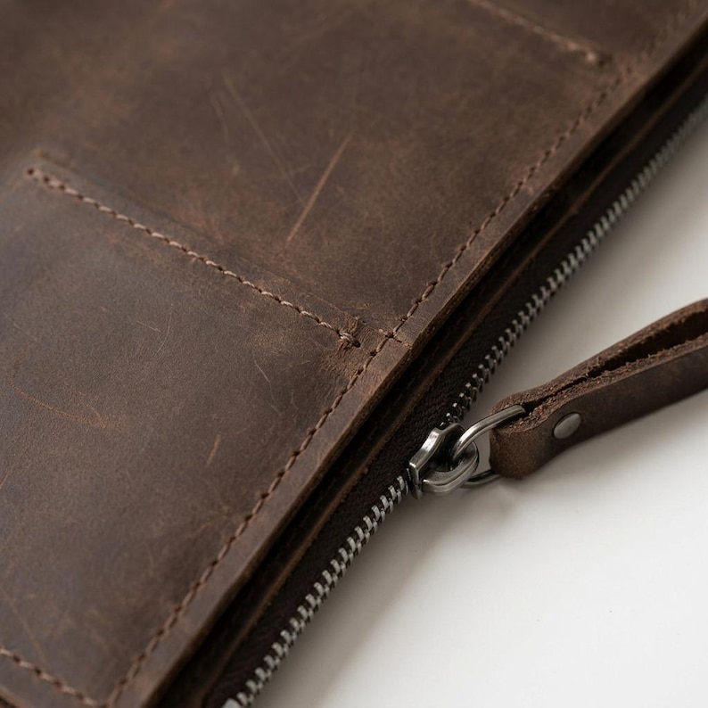 May include: A dark brown leather portfolio featuring a visible grain and natural surface scratches. The item includes a metal zipper closure with a matching leather pull tab and a stitched pocket detail on the exterior surface of the case.