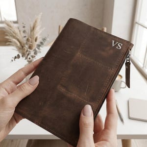 May include: A dark brown leather wallet featuring a smooth matte finish, a vertical zippered side compartment with a matching leather pull tab, and the embossed initials VS in the upper right corner. The wallet has visible stitching details.