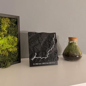 Könnte beinhalten: Eine schwarze Reliefkarte mit einer weißen Linie, die einen Weg darstellt. Die Karte trägt den Text "51.960 km 3.832 hm 20.71 h" am unteren Rand. Ein gerahmtes Mooskunstwerk und ein Glasterrarium mit Korken sind ebenfalls zu sehen.