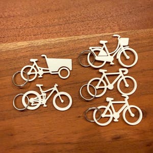 Könnte beinhalten: Eine Sammlung weißer Fahrrad-Schlüsselanhänger mit silbernen Schlüsselringen. Die Schlüsselanhänger zeigen verschiedene Fahrraddesigns, darunter ein Lastenrad und Fahrräder mit Körben. Die Schlüsselanhänger liegen auf einer Holzoberfläche.